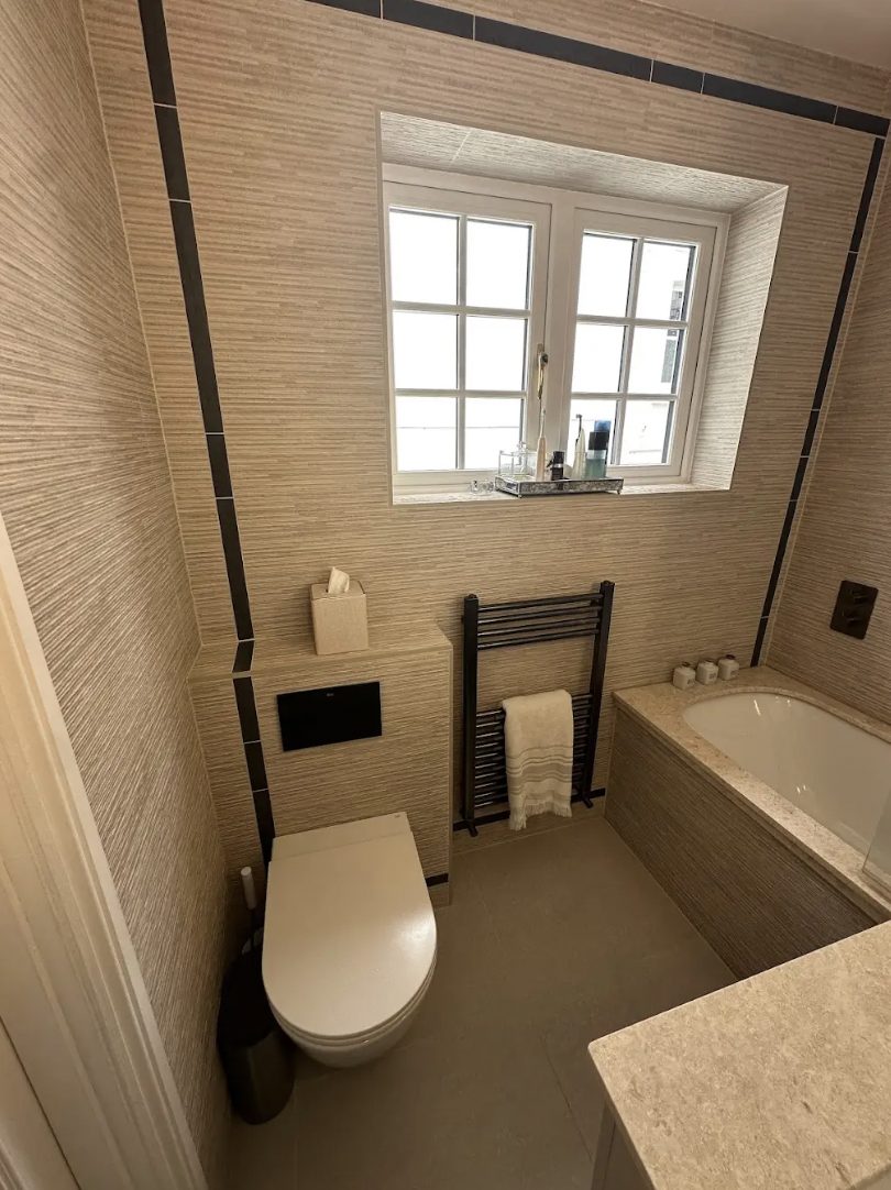 Grasscloth-effect ensuite with a wall-hung WC, black towel rail, window and a bath