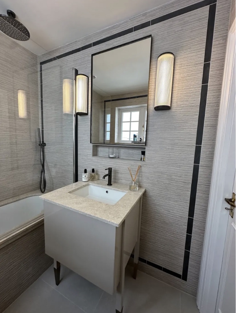 Cloakroom in grey grasscloth-effect tile with a fluted vanity, tall sconces and a bath with rain shower