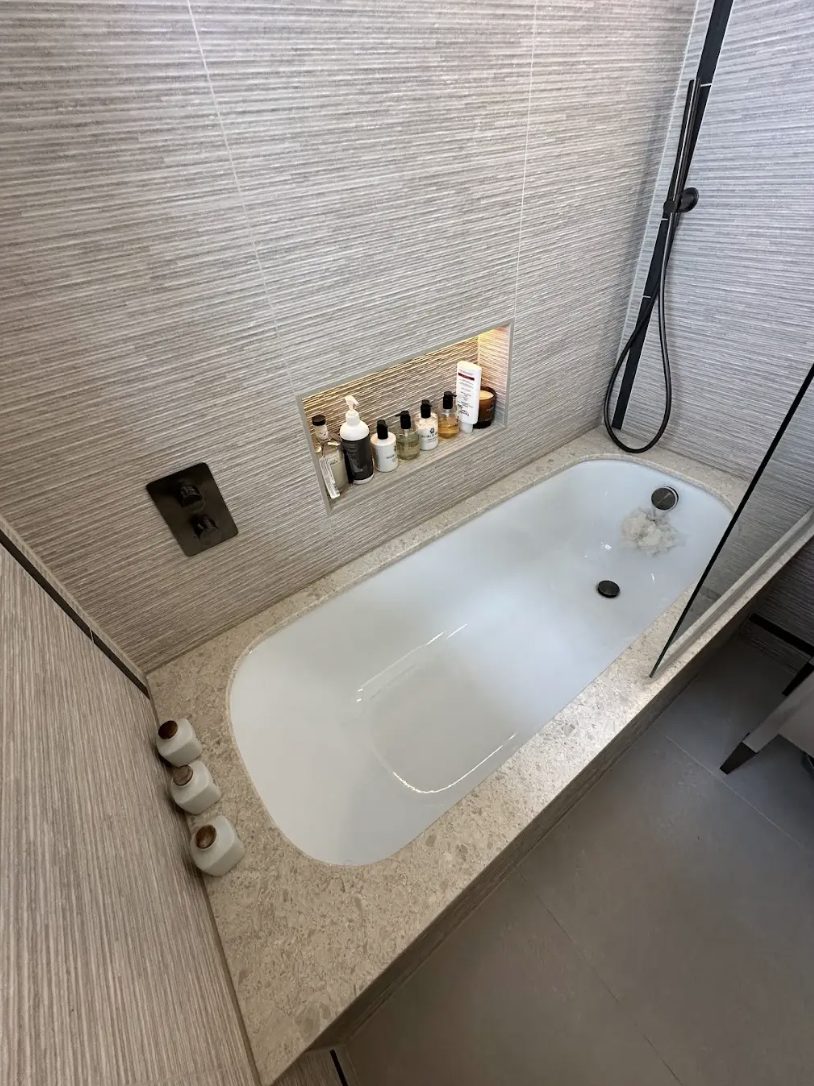 Inset bath in a grasscloth-effect bathroom with a lit recessed niche of bottles and a black filler