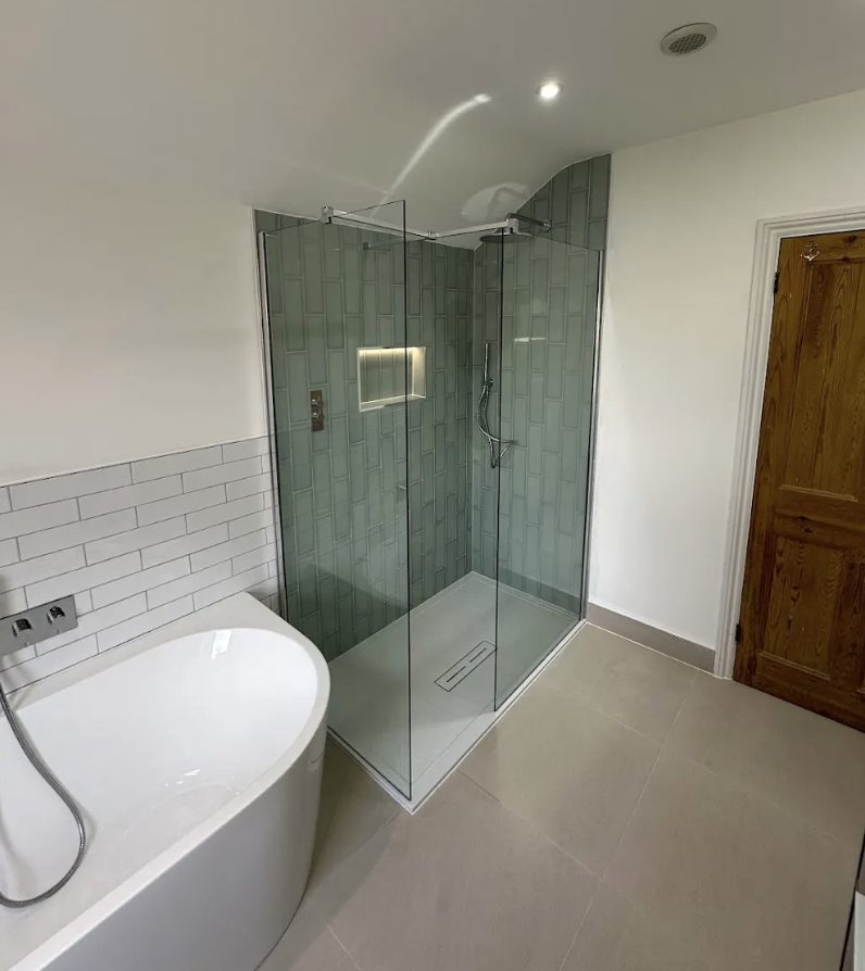Walk-in shower in sage-green metro tile beside a freestanding bath, white metro wainscot and a pine door