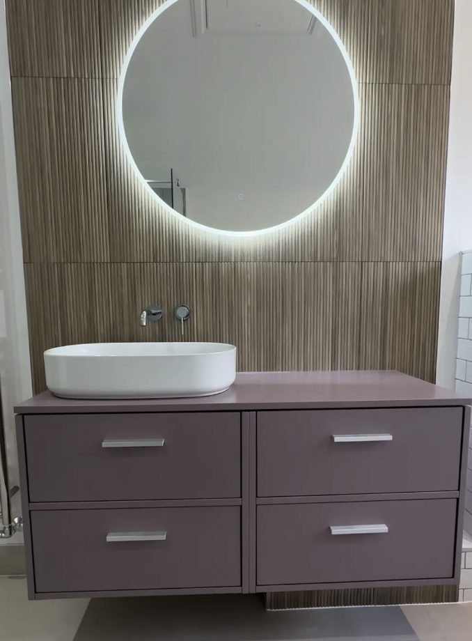 Vessel basin on a mauve four-drawer vanity below a round backlit mirror on a fluted tiled wall