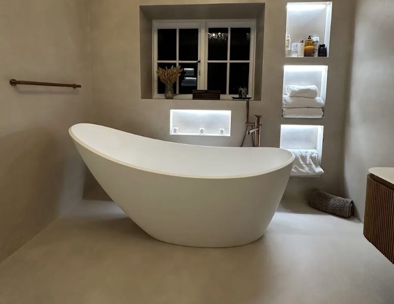 Stone slipper bath in a microcement bathroom with lit recessed niches holding folded towels