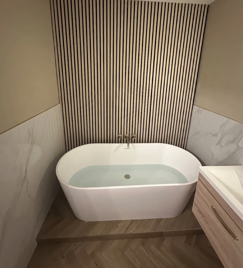 Freestanding oval bath against a full-height oak slat wall with marble wainscot and a herringbone floor