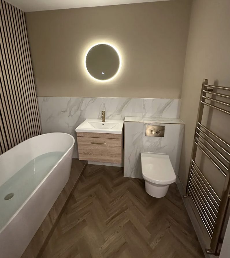 Freestanding bath below a round backlit mirror on an oak slat wall with marble tiling and herringbone floor