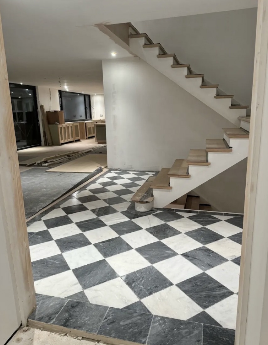 A period house mid-renovation: a new hardwood staircase above a black-and-white marble checkerboard hallway floor being laid