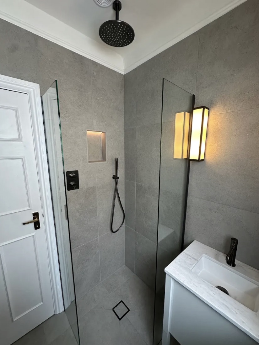 Walk-in shower room in grey stone tile with a black rain head, a backlit sconce and a compact vanity
