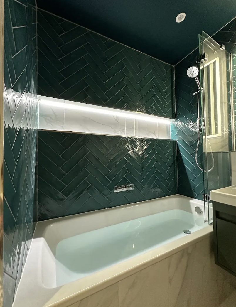 Bath with a teal herringbone tiled surround, a lit marble shelf and a walk-in shower
